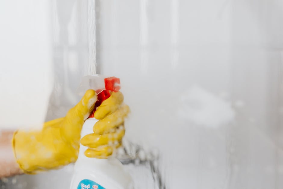 Close-up of a person's hand wearing a yellow rubber glove holding a white spray bottle with a red nozzle, applying cleaning solution to a glass shower door in a bathroom. The glass surface is wet and