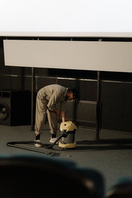 A professional cleaner dressed in full white protective gear, including a suit, gloves, and a face mask, is using a vacuum or extraction machine to deep clean a beige, textured carpet in a bright, min